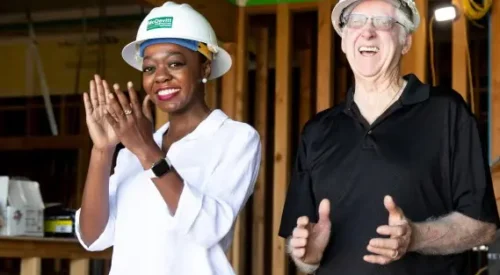 Woman and man in hard hats smiling and applauding in building under construction.