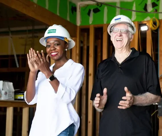 Woman and man in hard hats smiling and applauding in building under construction.