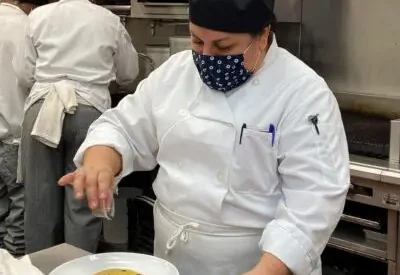 Trainee in white chef coat sprinkles garnish on plate of polenta