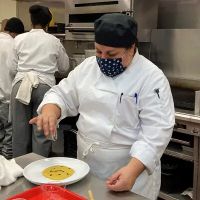 Trainee in white chef coat sprinkles garnish on plate of polenta