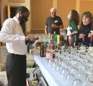 Man wearing white shirt and black trousers pours glass of wine for a guest at a bar holding glasses and bottles.