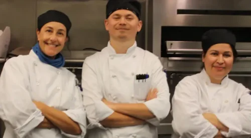 Three students in white chef's coats smiling in the kitchen with arms crossed