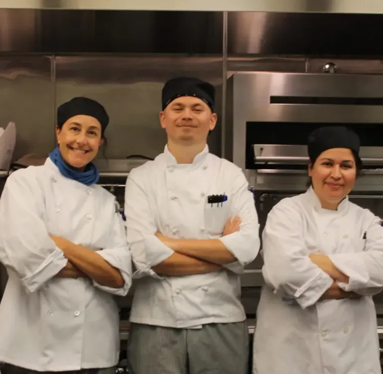 Three students in white chef's coats smiling in the kitchen with arms crossed