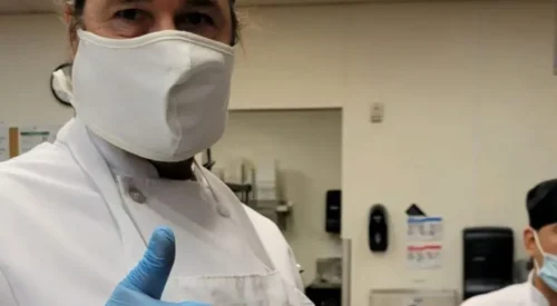 Chef trainee in white coat gives thumbs up in the kitchen. He wears a white mask but his eyes are smiling.