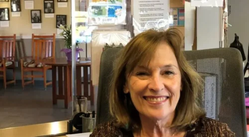 Woman smiles toward the camera wearing brown shirt and sweater. She is sitting in an office with a desk and reception window behind her.