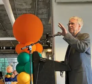 Man in gray blazer speaks at a lecture standing next to colorful balloons