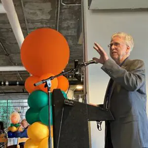 Man in gray blazer speaks at a lecture standing next to colorful balloons