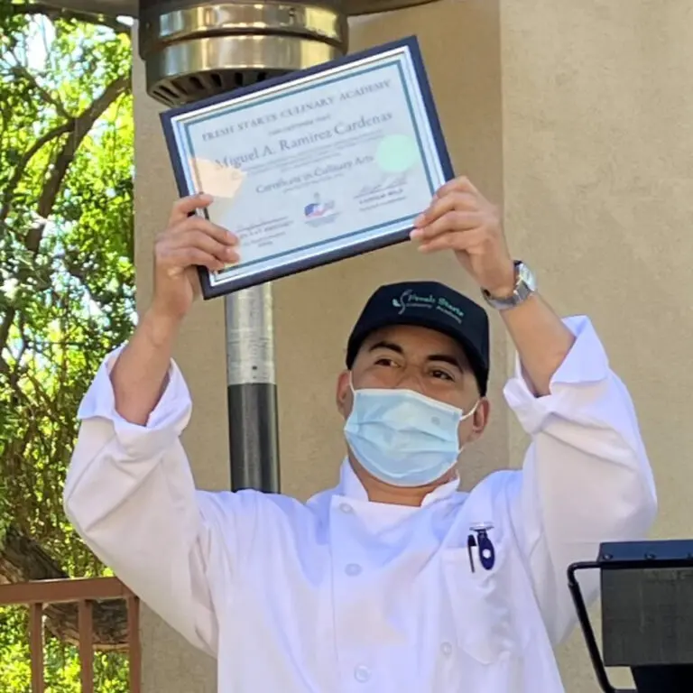 Young man in white chef's coat holds framed certificate over his head.