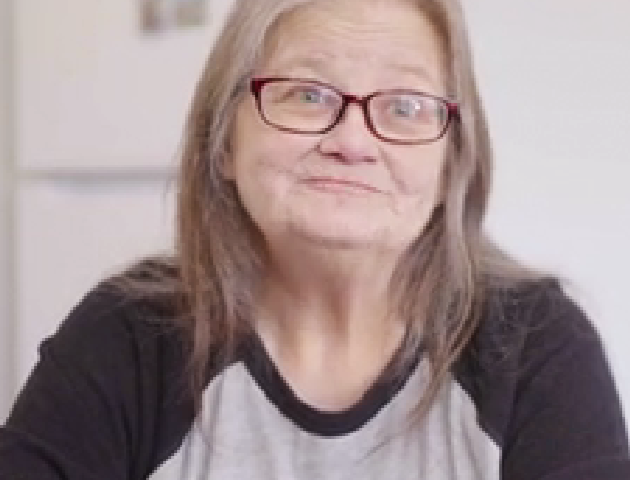 Woman with gray hair and glasses smiles at camera. She is wearing gray and black T-shirt.
