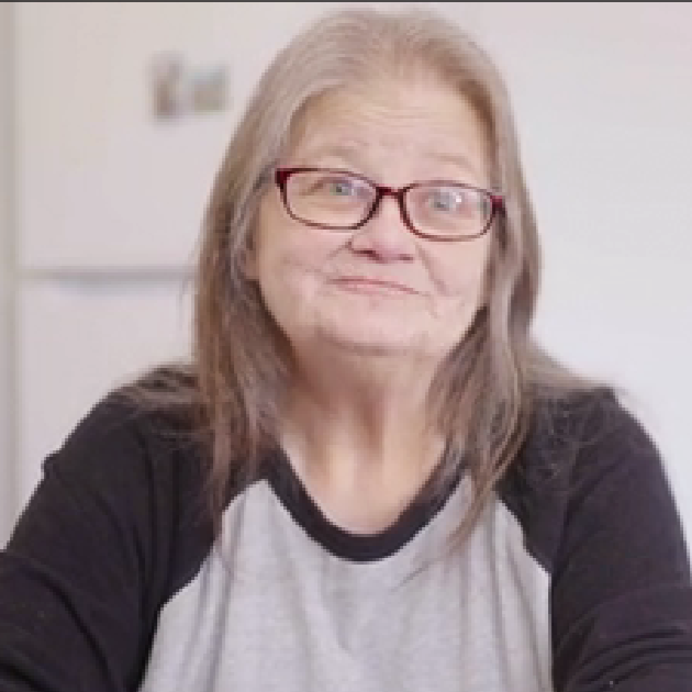 Woman with gray hair and glasses smiles at camera. She is wearing gray and black T-shirt.