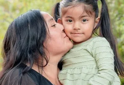 A mother holder a small child, kissing her on the cheek.