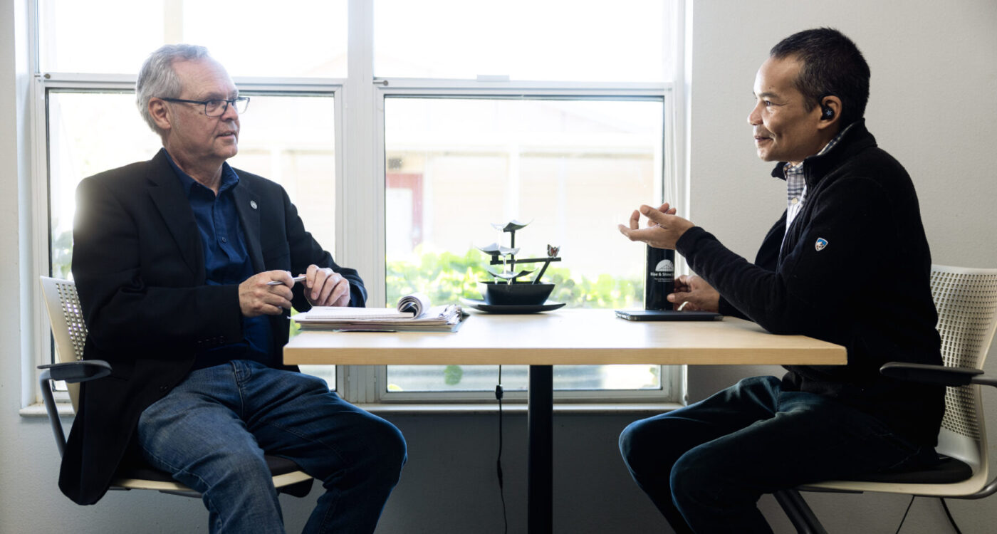 two men sit across a table having a conversation.