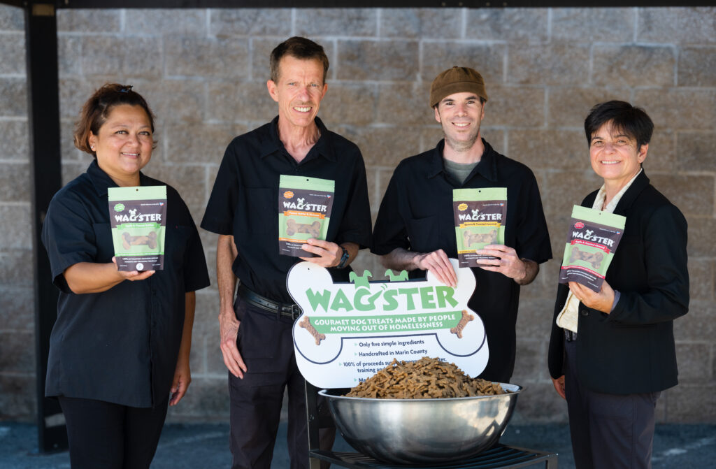 Four members of the wagster team holding bags of treats and standing in front of sign reading 'Wagster, gourmet dog treats made by people moving out of homelessness'
