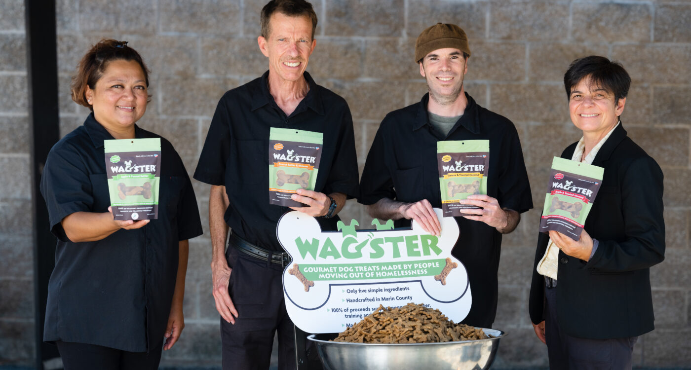 Four members of the wagster team holding bags of treats and standing in front of sign reading 'Wagster, gourmet dog treats made by people moving out of homelessness'