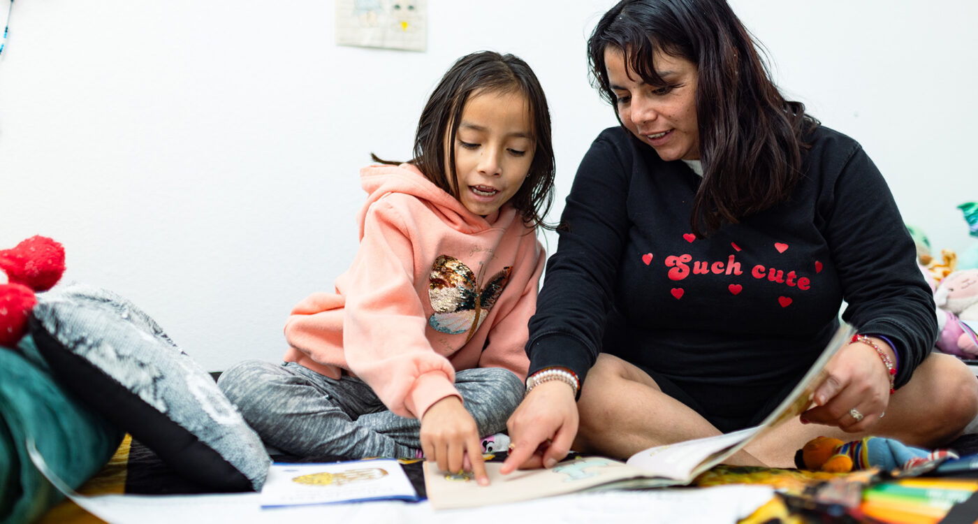 a mom and daughter sit on a bed reading from a storybook