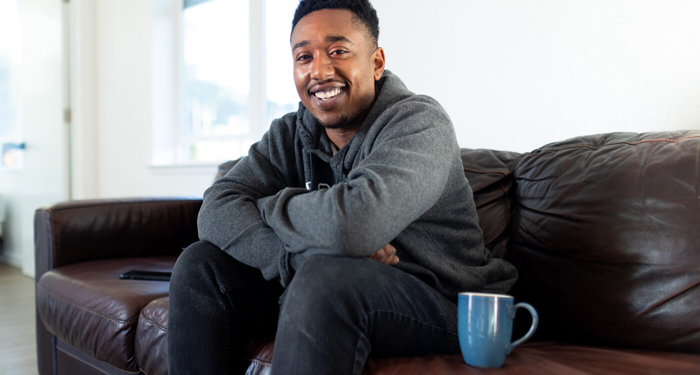 a man sits on a sofa smiling to the camera with a coffee mug next to him