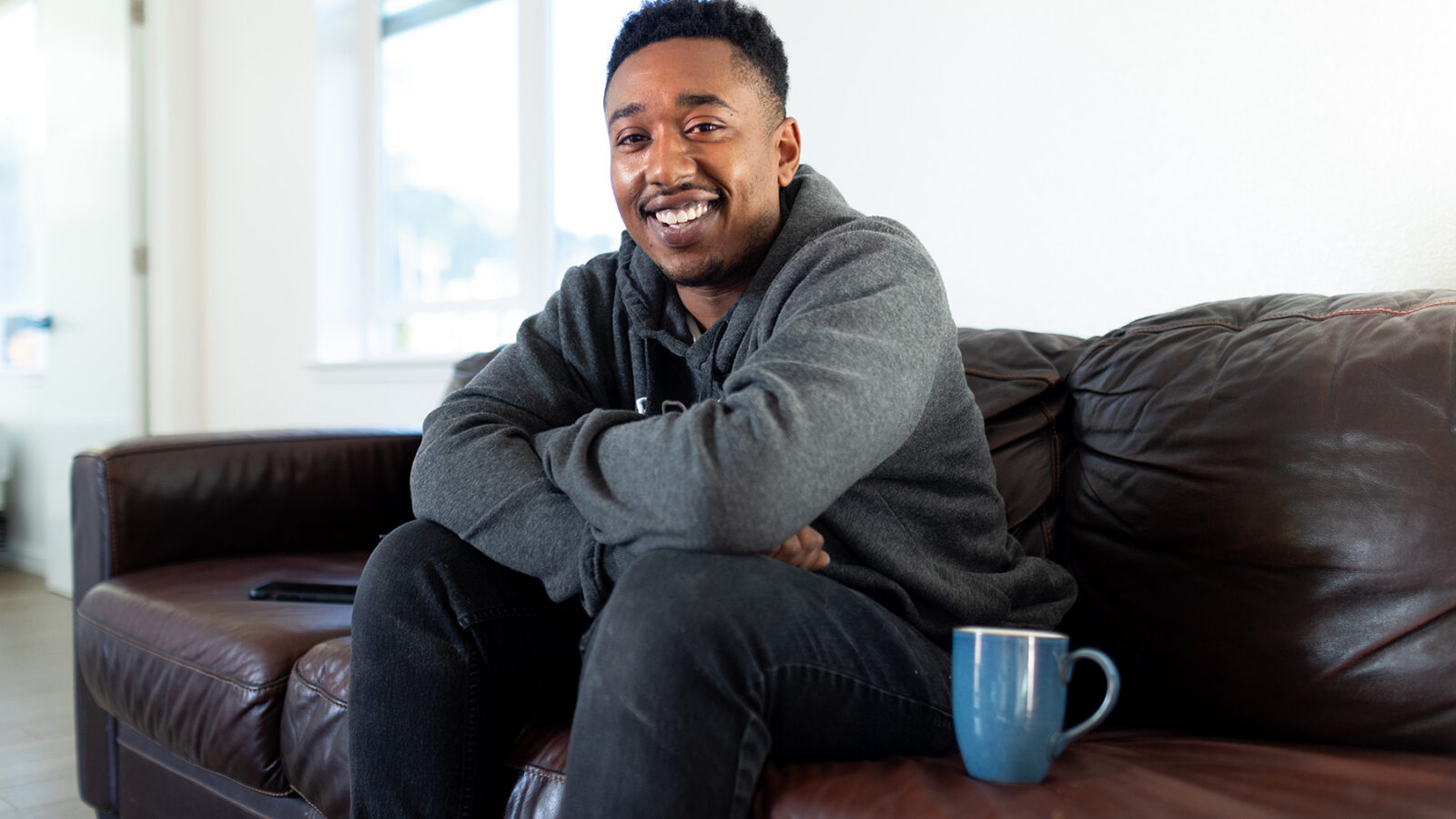 a man sits on a sofa smiling to the camera with a coffee mug next to him
