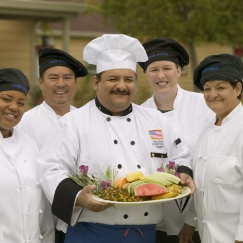 Five chefs posing for the camera, one holding a tray of food