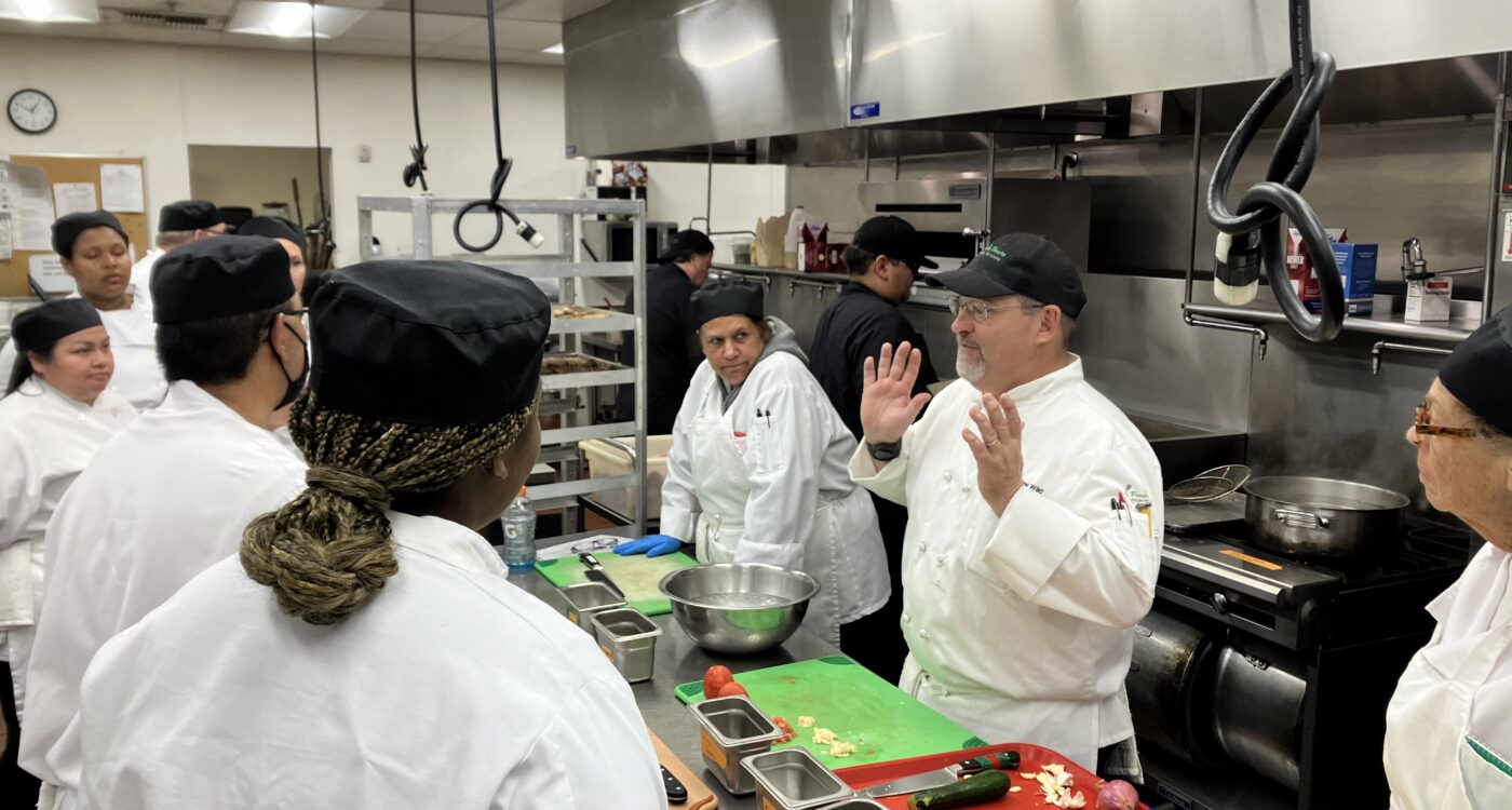 Four chefs in a kitchen. One is talking.