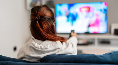 a person, facing away from the camera, sits on a sofa pointing a remote at the television.