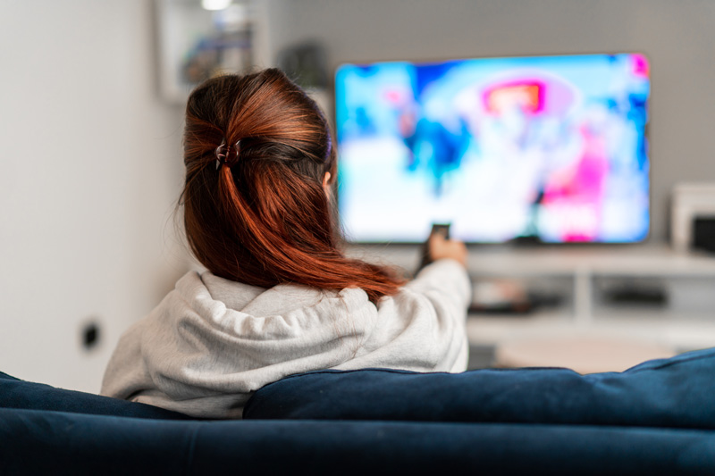 a person, facing away from the camera, sits on a sofa pointing a remote at the television.