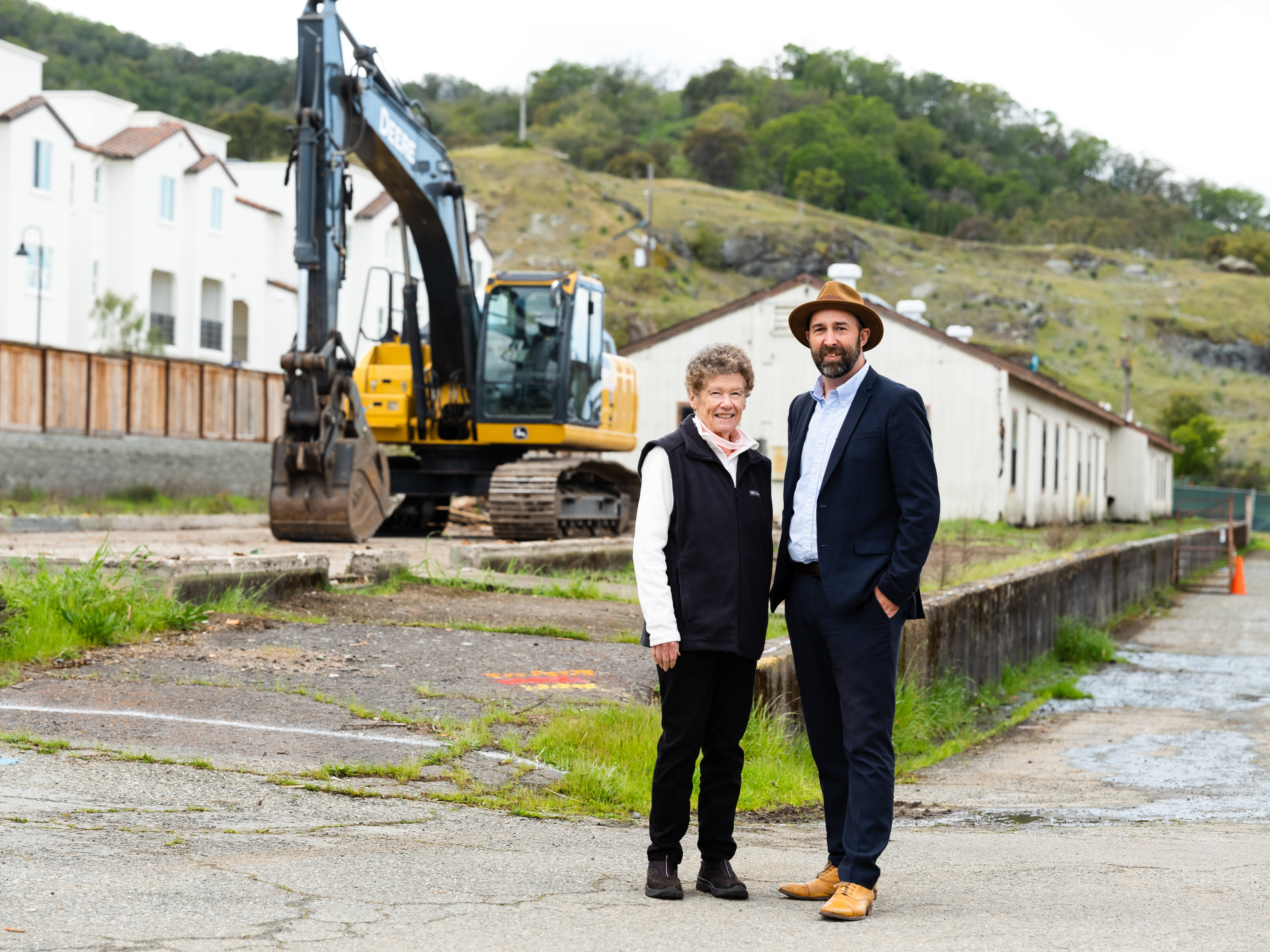 Mary Kay Sweeney and Paul Fordham stand in front of a construction site with a backhoe behind them