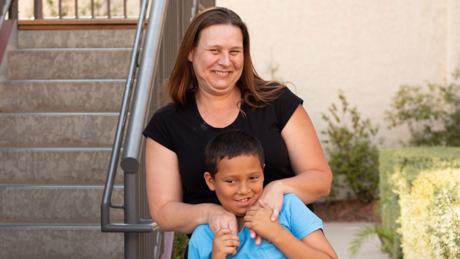 A mom stands behind her son next to a staircase