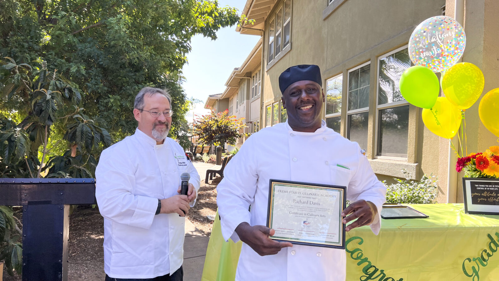 A man holds up his certificate of culinary arts after completing the fresh starts culinary academy. His teacher stands next to him.