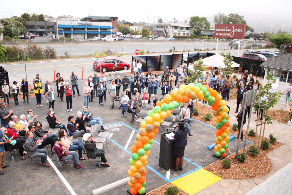 people gathered in a parking lot while a speech is given under a balloon archway