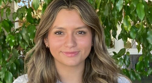 Teenage girl smiles at the camera. She has long curly light-brown hair and wears a white sweater.