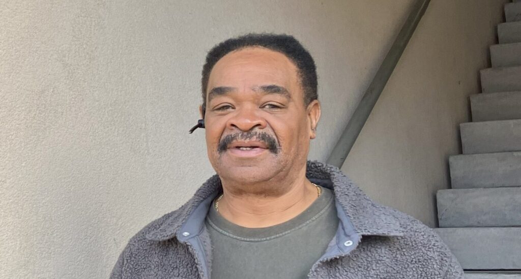 Black man with short hair smiles at the camera. He is wearing gray shirt and standing by an apartment staircase.