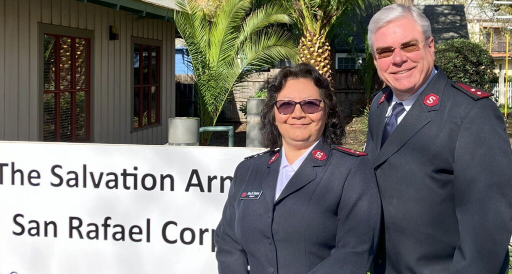 Dark-haired woman in sunglasses and taller gray-haired man stand by sign that says 'The Salvation Army - San Rafael Corps.' They wear gray Salvation Army jackets.