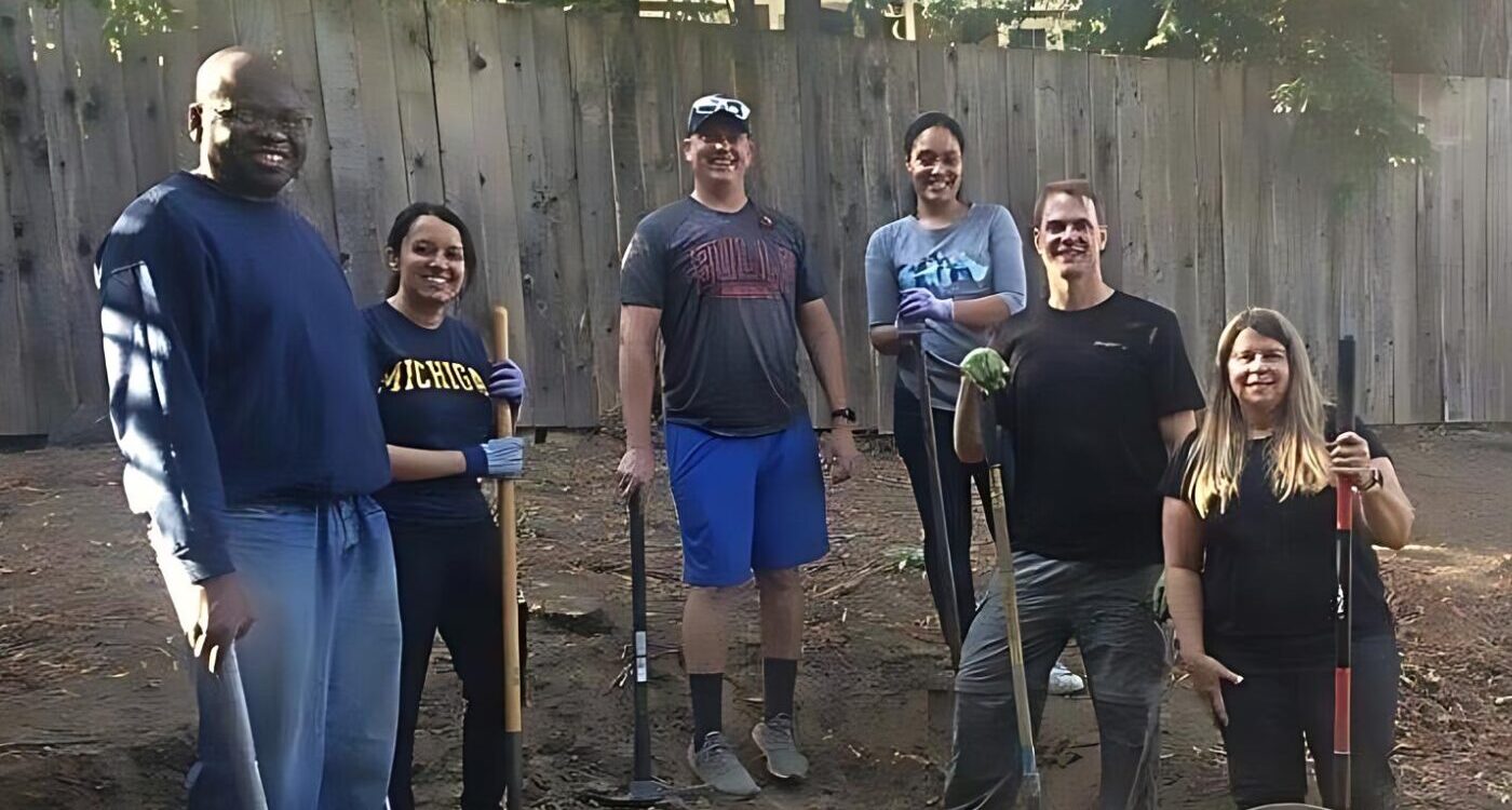Six volunteers in T-shirts and jeans smile at the camera. They are standing in a backyard garden holding rakes and other tools.