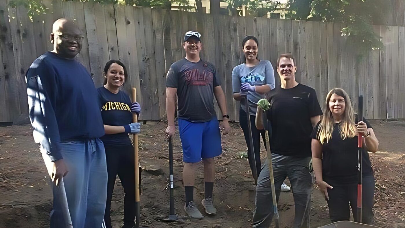 Six volunteers in T-shirts and jeans smile at the camera. They are standing in a backyard garden holding rakes and other tools.