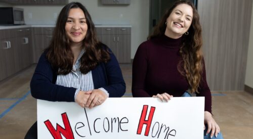 Two women in a brand new apartment kneel with a large sign that says "Welcome Home"