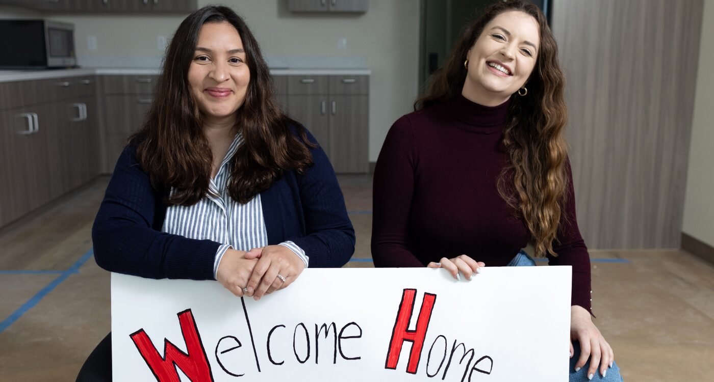 Two women in a brand new apartment kneel with a large sign that says "Welcome Home"