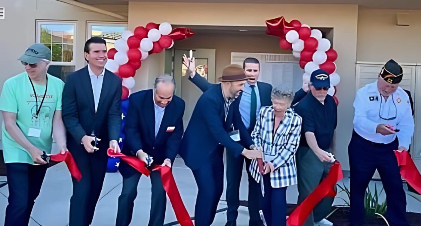 Eight people holding scissors smile as they cut red ribbon to symbolically open the new veterans housing.