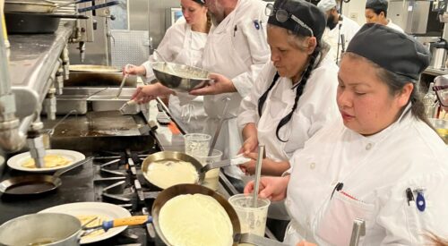 Culinary students wearing white coats and black caps stand at stove making crepes.