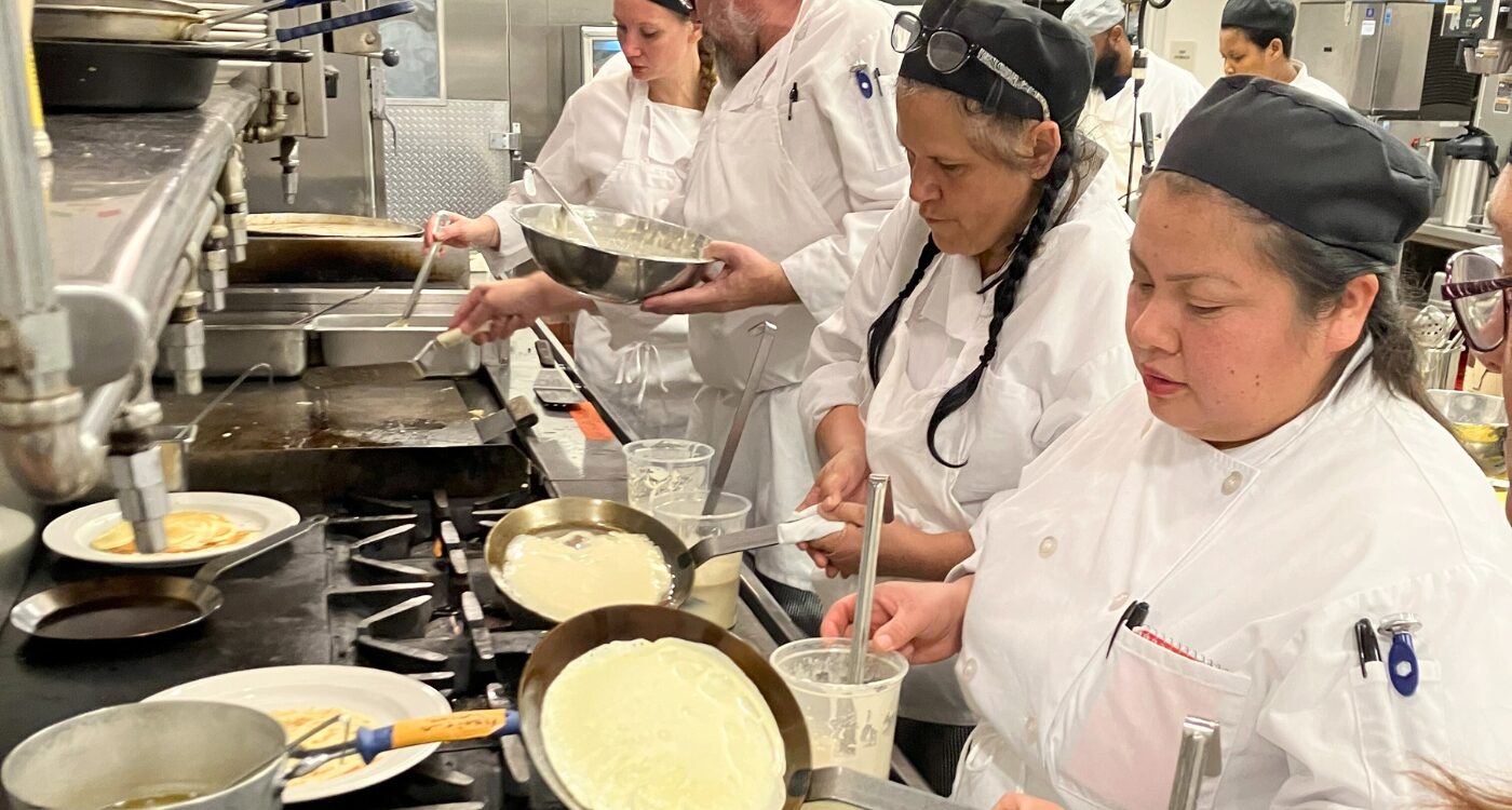 Culinary students wearing white coats and black caps stand at stove making crepes.