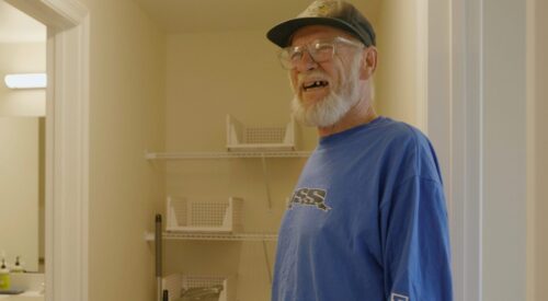 Light-skinned man with glasses and white beard is smiling while standing in new apartment.