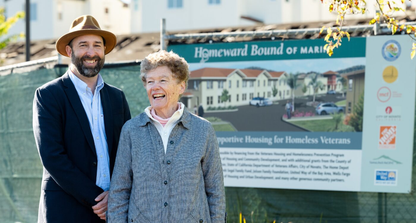Man in dark suit with hat and woman in gray shirt stand near construction sign.