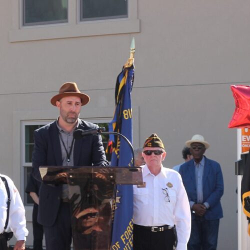 Man in brown hat and blue suit addresses crowd from the lectern.