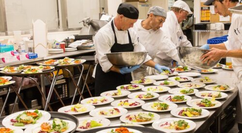 Four men in chef's coats hold bowls of salad that they are arranging on plates lined up on a steel table.