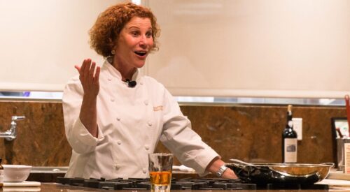 Chef with curly red hair talks to the audience. She is wearing a white chef's coat.