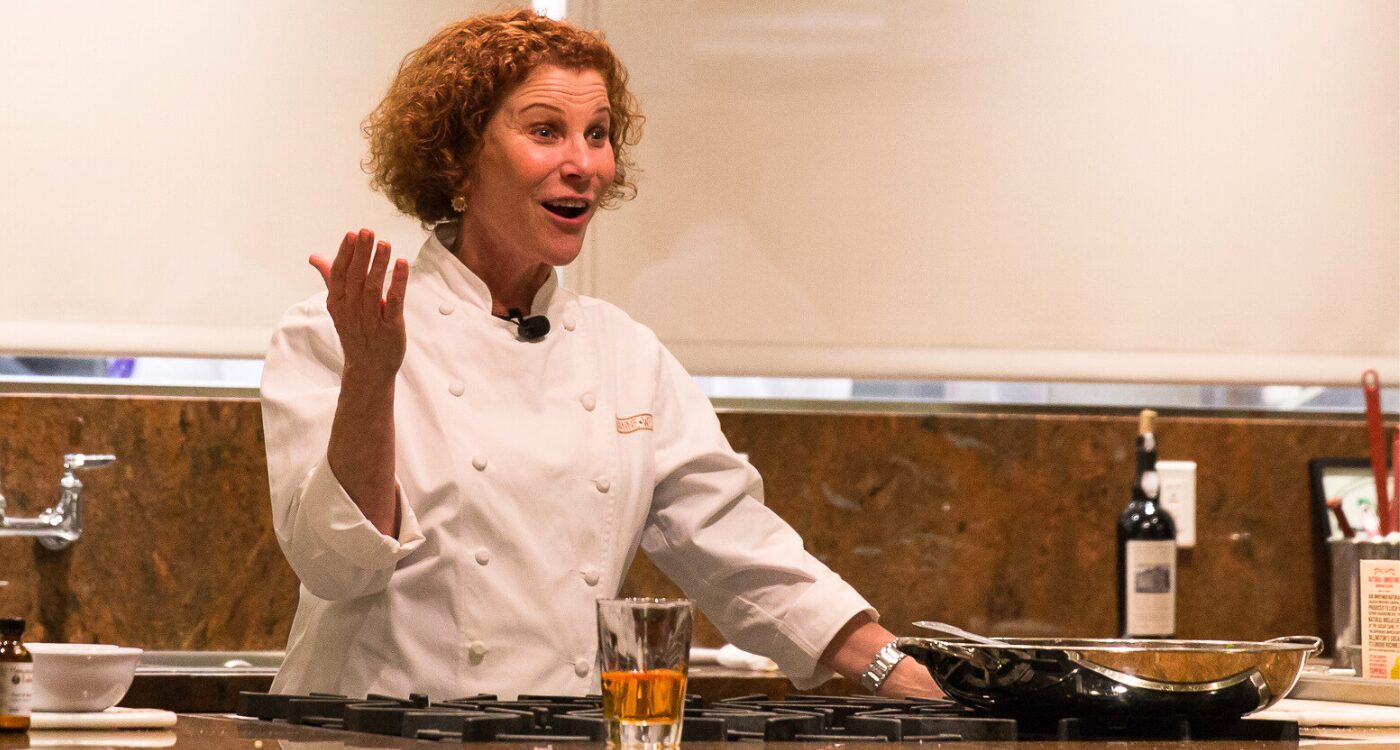 Chef with curly red hair talks to the audience. She is wearing a white chef's coat.