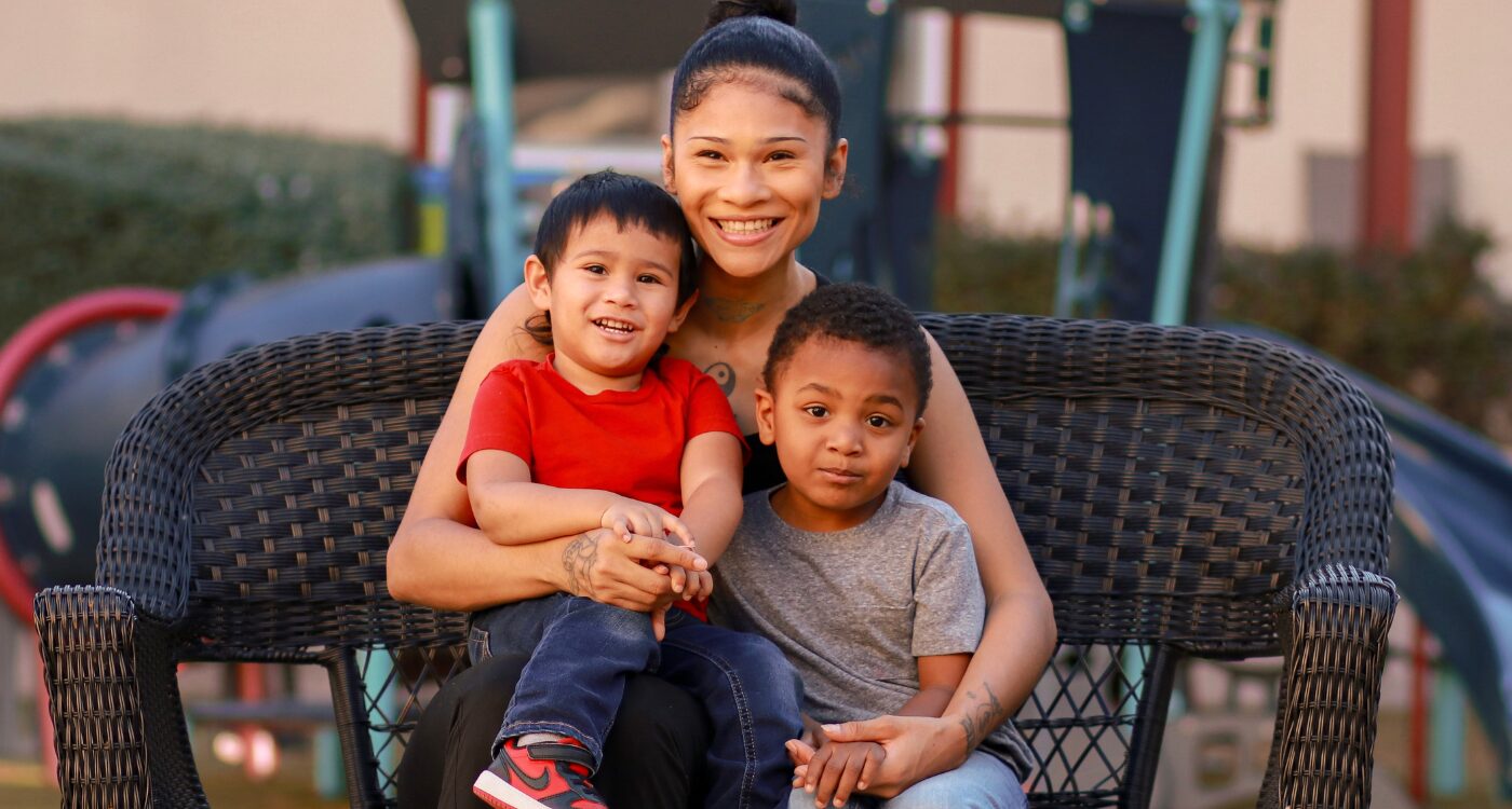 Woman with dark hair pulled back smiles at the camera. She has her arms around two young boys leaning on her.