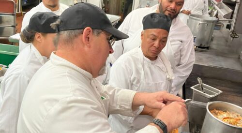 Chef instructor points to his spoon as he works at the stove with culinary students.