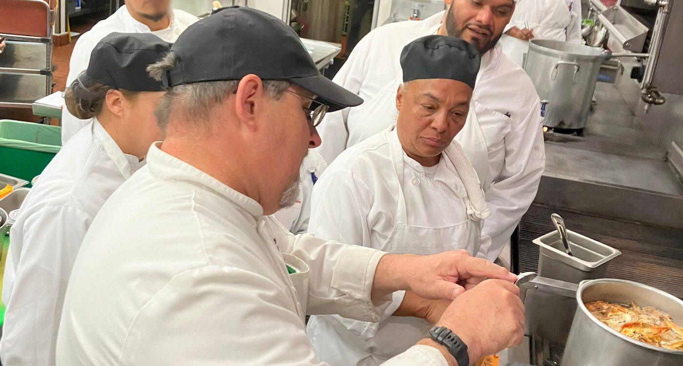 Chef instructor points to his spoon as he works at the stove with culinary students.