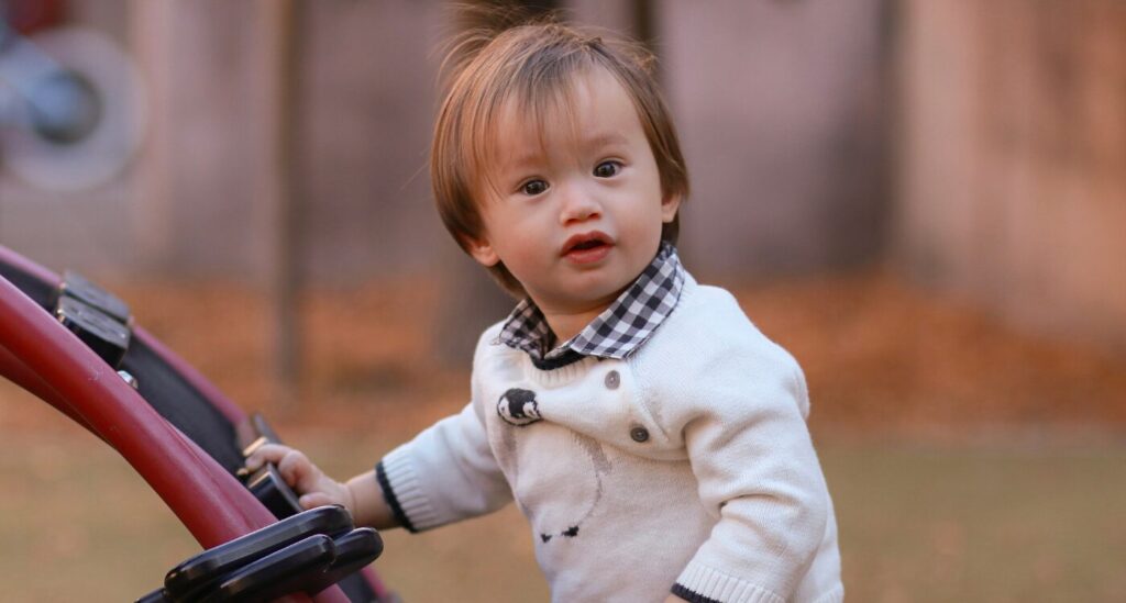 Toddler boy in white sweater stands in the playground and looks at the camera