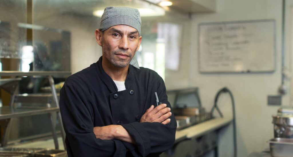 Man wearing black and white checkered hat and black chef's coat stands in the kitchen. He looks at the camera with arms folded.